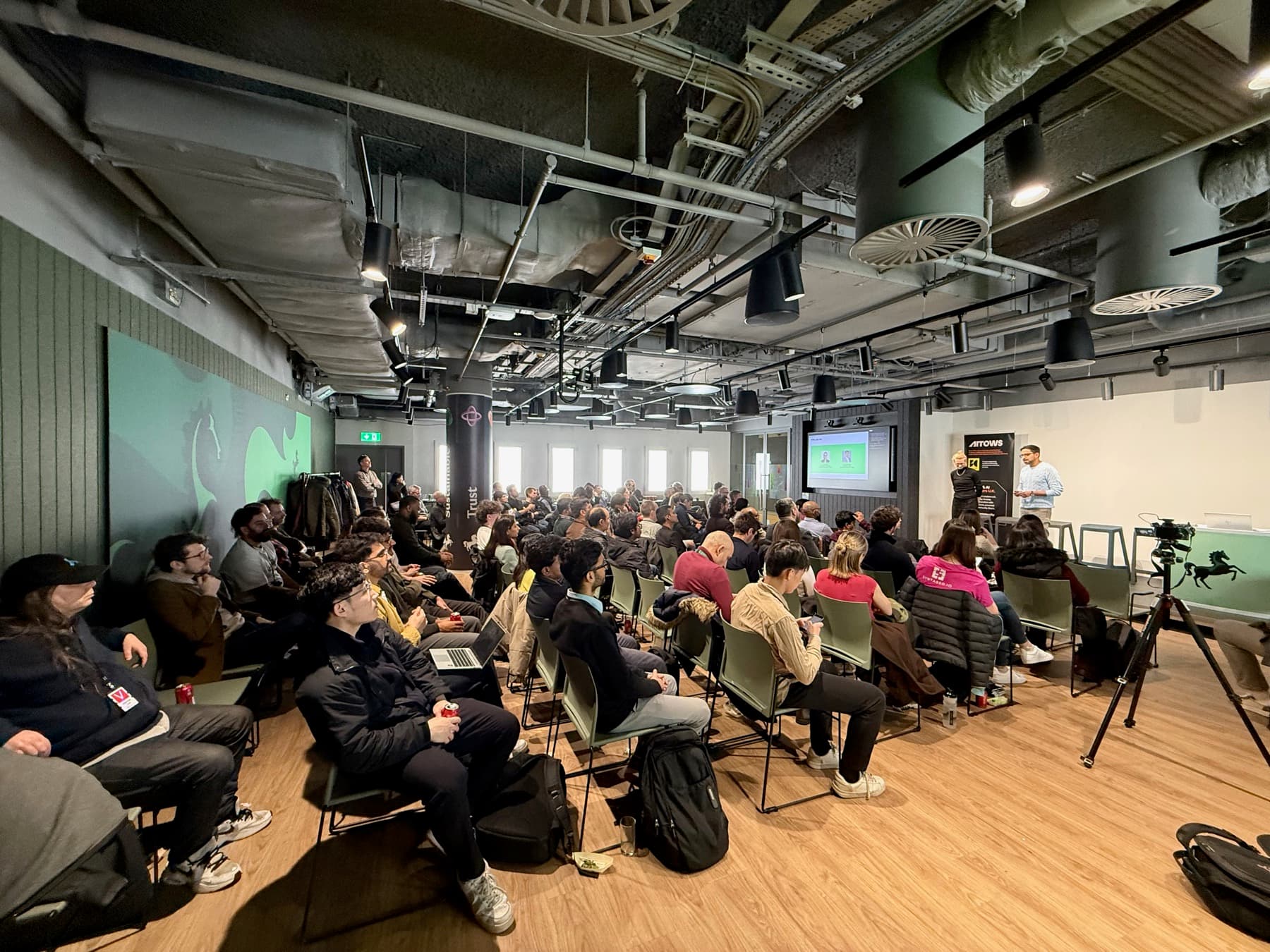 A full hall of attendees watching a talk under an industrial ceiling